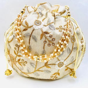 May include: Cream-colored drawstring bag with gold and silver floral embroidery. The bag features a beaded handle and gold accents on the drawstring and base. The floral design includes white flowers and gold leaves.