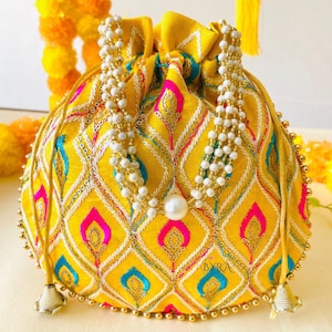 May include: A yellow drawstring pouch with an intricate embroidered pattern in pink, blue, and white. The bag is adorned with white beaded strands and gold accents. The pouch has a pearl at the center and gold tassels.