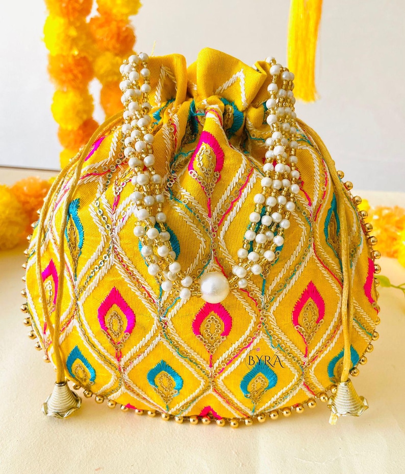 May include: A yellow drawstring pouch with an intricate embroidered pattern in pink, blue, and white. The bag is adorned with white beaded strands and gold accents. The pouch has a pearl at the center and gold tassels.
