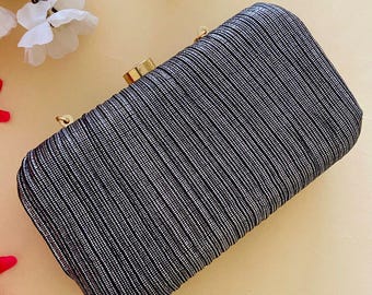 May include: A rectangular clutch with a textured, vertically striped pattern in black and silver. The clutch has a gold-colored clasp and hardware. The background is a light beige color, with white flowers and red petals in the corners.