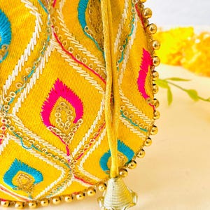 May include: A yellow, round embroidered bag with a drawstring closure. The bag features a geometric pattern in pink, blue, and white thread, accented with gold sequins and small gold beads along the edge. A gold tassel hangs from the drawstring.