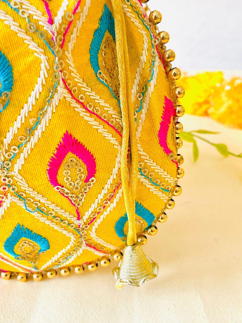 May include: A yellow, round embroidered bag with a drawstring closure. The bag features a geometric pattern in pink, blue, and white thread, accented with gold sequins and small gold beads along the edge. A gold tassel hangs from the drawstring.
