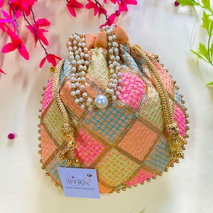 May include: An embroidered pouch with a drawstring closure, featuring a geometric pattern in pink, orange, and blue, with gold trim. A pearl necklace and a tag with the text "BYRA" are also visible. The bag is a vibrant accessory.