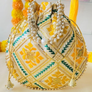 May include: A round, cream-colored pouch with yellow floral embroidery and green accents. The bag features a beaded drawstring and a pearl accent. Gold trim and a beaded edge complete the design. The text "BYRA" is visible.