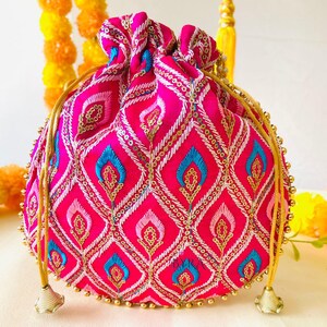 May include: A vibrant pink drawstring pouch with an intricate embroidered pattern. The design features diamond shapes with blue and gold accents, embellished with sequins. Gold beaded trim and drawstrings complete the look.