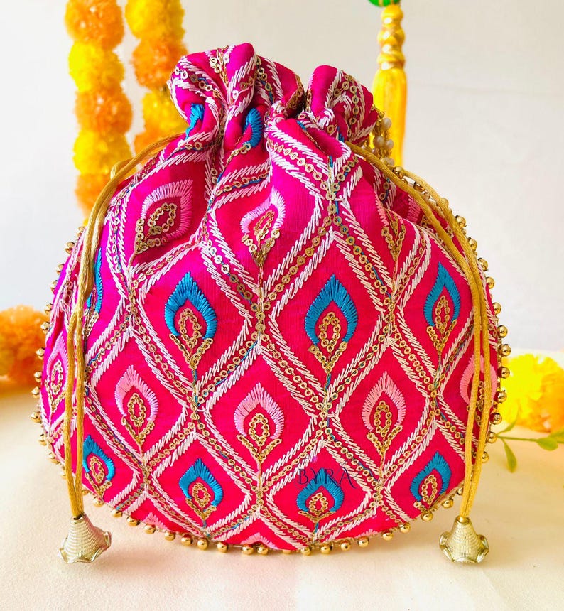 May include: A vibrant pink drawstring pouch with an intricate embroidered pattern. The design features diamond shapes with blue and gold accents, embellished with sequins. Gold beaded trim and drawstrings complete the look.