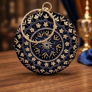 Può includere: Pochette rotonda nera con ricami dorati e blu. La pochette presenta un manico circolare ornato di scintillanti abbellimenti. Il design include motivi floreali e a stella, creando un accessorio elegante.