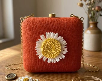 Könnte beinhalten: Eine orangefarbene quadratische Handtasche mit goldener Kette und einer großen Gänseblümchen-Applikation. Das Gänseblümchen hat eine gelbe Mitte und weiße Blütenblätter. Auf einer Holzoberfläche liegen Gänseblümchen und ein Knopf.