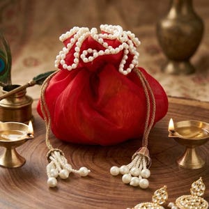 Peut inclure: Une pochette rouge à cordon de serrage avec une bordure et des glands perlés blancs. La pochette est faite d'un tissu transparent et possède des cordons dorés. Les glands sont ornés de perles blanches. La pochette est probablement utilisée pour des bijoux ou de petits cadeaux.