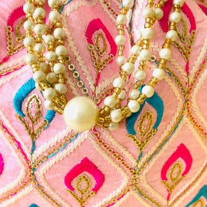 May include: A pink embroidered pouch with a beaded necklace. The pouch features a diamond pattern with gold, blue, and fuchsia accents. The necklace has multiple strands of white and gold beads, with a large pearl at the bottom.