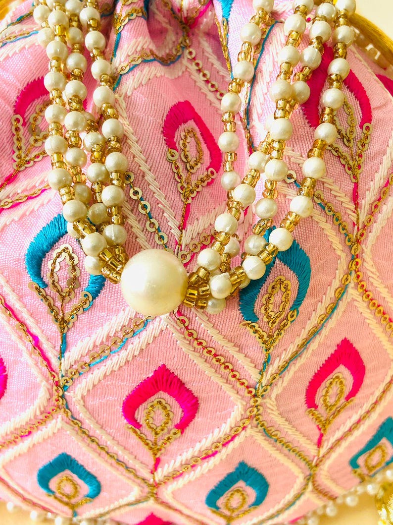 May include: A pink embroidered pouch with a beaded necklace. The pouch features a diamond pattern with gold, blue, and fuchsia accents. The necklace has multiple strands of white and gold beads, with a large pearl at the bottom.