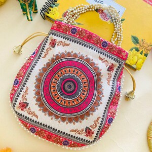 May include: A white and pink drawstring bag with a detailed mandala design. The bag has a pearl beaded handle and gold-coloured drawstring cords. The edges are trimmed with small white beads. The bag is decorated with floral and paisley patterns.