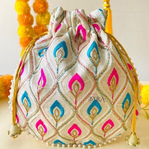 May include: Cream-colored drawstring pouch with a repeating pattern of blue and pink teardrop shapes. The design is embroidered with gold thread and sequins. The bag has gold drawstrings and tassels, and is trimmed with small white beads. The word "BYRA" is visible on the bag.
