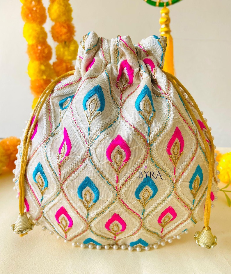 May include: Cream-colored drawstring pouch with a repeating pattern of blue and pink teardrop shapes. The design is embroidered with gold thread and sequins. The bag has gold drawstrings and tassels, and is trimmed with small white beads. The word "BYRA" is visible on the bag.