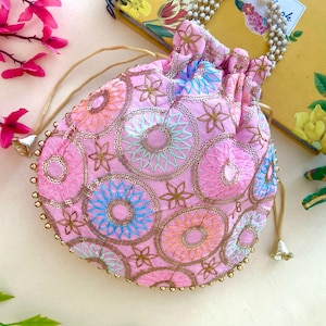 May include: A pink drawstring pouch with embroidered circular and floral patterns in shades of pink, blue, orange, and gold. The pouch has a gold beaded trim and a pearl-beaded handle. The drawstring is a light tan colour.