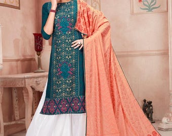 May include: A teal knee-length tunic featuring a dense gold and magenta floral pattern, paired with solid white wide-leg trousers that have a gold border at the hem. A large peach-colored shawl with a repeating floral print drapes over the shoulder.