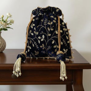 Può includere: Una borsa a coulisse in velluto blu navy con ricami floreali dorati e paillettes. La borsa presenta un cordoncino dorato, nappe di perline e un bordo decorativo. Questa elegante borsa è adatta per occasioni speciali.