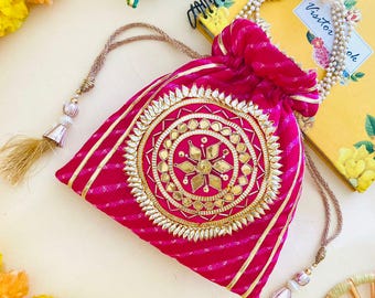 May include: A vibrant pink drawstring bag with gold embellishments and a detailed circular design. The bag features a striped pattern and gold tassels. A small green elephant figurine and a yellow notebook are in the background.
