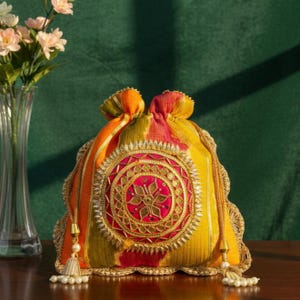 Puede incluir: Un bolso con cordón amarillo, naranja y rojo con intrincados bordados dorados y un diseño circular central. El bolso presenta un borde festoneado y detalles de borlas, con un cierre de cordón con cuentas. El fondo es verde oscuro.