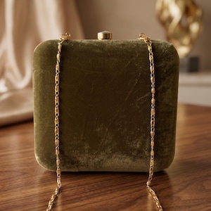 May include: An olive green velvet clutch bag with a gold chain strap. The rectangular bag has rounded corners and a small clasp. The gold chain is delicate and hangs down on either side of the bag. The clutch sits on a wooden surface.