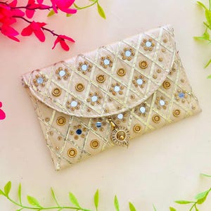 May include: A beige clutch with a quilted pattern, embellished with gold-colored embroidery, small mirrors, and decorative gold accents. The clutch has a flap closure and a gold-colored tassel. The background includes pink flowers and green leaves.