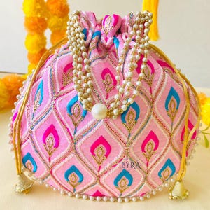 May include: Pink embroidered pouch with a drawstring closure and a beaded handle. The pouch features a repeating pattern of pink, blue, and gold shapes. The pouch is adorned with a pearl beaded trim and a large pearl at the center. The text "BYRA" is visible.