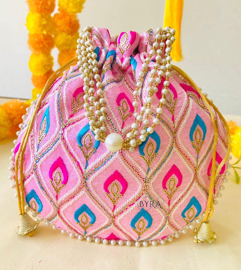 May include: Pink embroidered pouch with a drawstring closure and a beaded handle. The pouch features a repeating pattern of pink, blue, and gold shapes. The pouch is adorned with a pearl beaded trim and a large pearl at the center. The text "BYRA" is visible.