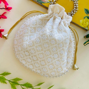 May include: Cream-coloured, embroidered drawstring bag with a floral pattern. The bag has a beaded handle and a gold-coloured drawstring. The bag is decorated with small white beads around the bottom edge. A yellow notebook with the words "Visitor Book" is in the background.