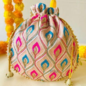 May include: A peach-colored drawstring pouch with an embroidered pattern of blue and pink teardrop shapes. The pouch has gold-colored beaded trim and drawstrings. The text "BYRA" is visible on the lower right side of the pouch.