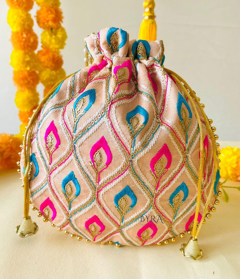 May include: A peach-colored drawstring pouch with an embroidered pattern of blue and pink teardrop shapes. The pouch has gold-colored beaded trim and drawstrings. The text "BYRA" is visible on the lower right side of the pouch.