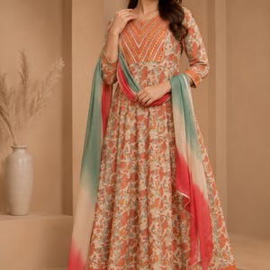 Puede incluir: Un vestido largo de color beige con estampado floral naranja y verde, con mangas tres cuartos, cuello redondo con bordado naranja y un dupatta degradado a juego en tonos verde y coral, combinado con pantalones de color naranja sólido.