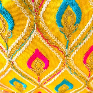 May include: Close-up of a yellow fabric with intricate embroidery. The design features blue, pink, and white stitching, along with gold sequins. The pattern includes diamond shapes and stylized leaf motifs, creating a vibrant and detailed textile.