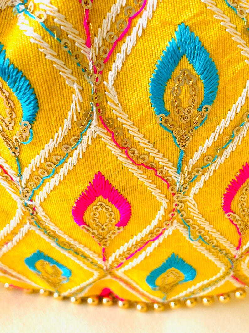 May include: Close-up of a yellow fabric with intricate embroidery. The design features blue, pink, and white stitching, along with gold sequins. The pattern includes diamond shapes and stylized leaf motifs, creating a vibrant and detailed textile.
