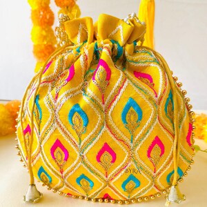 May include: A yellow drawstring pouch with intricate embroidery in a diamond pattern. The design incorporates pink, blue, and white accents, with gold sequin details. The pouch has a beaded trim and the word "BYRA" is visible.