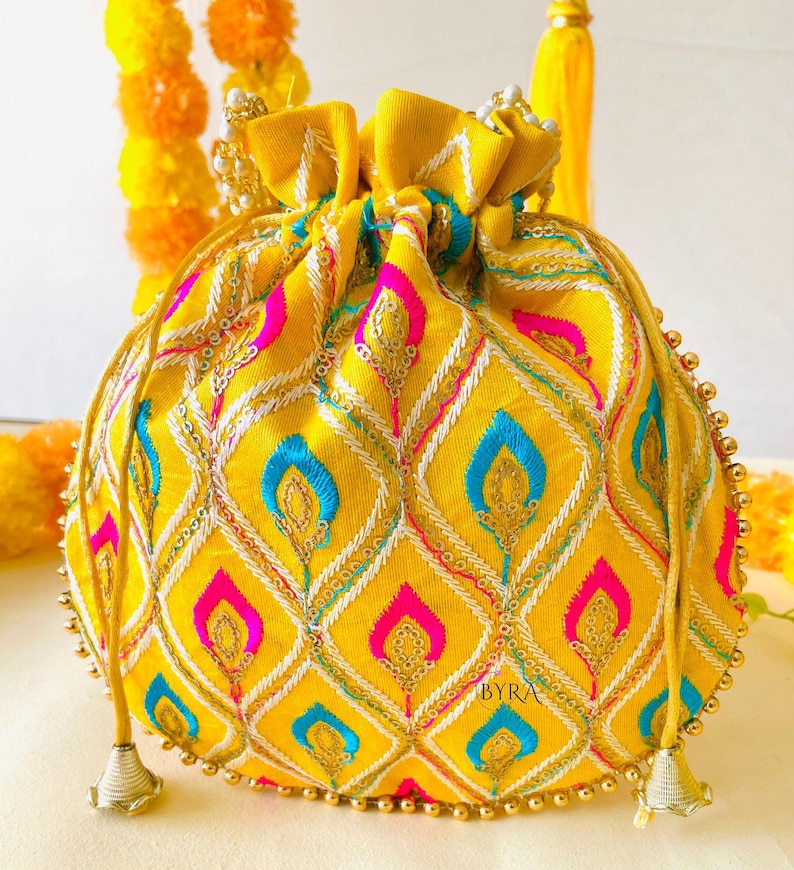 May include: A yellow drawstring pouch with intricate embroidery in a diamond pattern. The design incorporates pink, blue, and white accents, with gold sequin details. The pouch has a beaded trim and the word "BYRA" is visible.