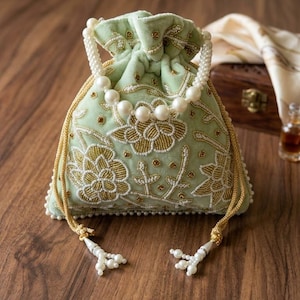 Peut inclure: Un sac à cordon en velours vert clair avec broderies florales dorées et blanches. Le sac est orné d'une poignée perlée et d'un cordon doré avec des perles. Le sac est posé sur une surface en bois.