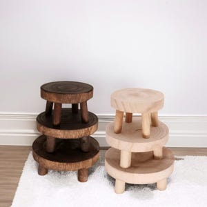 May include: Two wooden step stools. One is stained a dark brown, the other is natural wood. Each stool has three circular tiers supported by short, rounded legs. The stools are displayed on a white rug against a white wall.