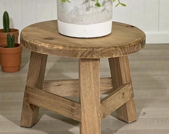 May include: A small, round wooden plant stand with a natural finish. The stand has three angled legs and a circular top. A potted plant sits on top of the stand. The stand is approximately 12 inches in diameter.