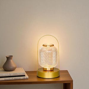 Puede incluir: Una lámpara de mesa dorada con una pantalla de cristal transparente con diseño de diamantes. La lámpara tiene una base redonda y un asa curvada. Un pequeño jarrón marrón y libros están sobre la mesa de madera.