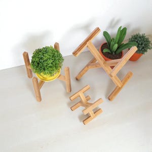 Op de afbeelding: Verschillende bamboe plantenstandaards in verschillende stadia van montage. Eén standaard houdt een gele pot met een groene plant vast. De standaards zijn gemaakt van lichtgekleurd bamboe en zijn ontworpen om potplanten te verhogen. Voor binnen of buiten.