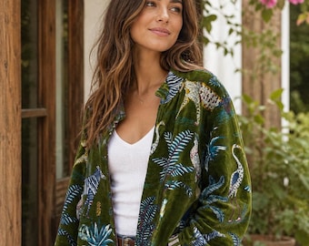 May include: A long-sleeved olive green velvet jacket featuring a vibrant jungle-inspired print with zebras, leopards, and tropical foliage. The jacket has a relaxed fit, an open front, and striped detailing on the inner cuffs, worn over a white top.