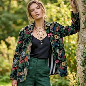 Pode incluir: Um blazer preto com uma estampa colorida de flores e animais, uma regata preta, calças largas verde-escuras e uma bolsa de couro marrom. O blazer possui um forro listrado em preto e branco e é usado com um colar dourado de várias camadas.