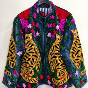 Op de afbeelding: Een blauw fluwelen jasje met geborduurde tijger- en bloemmotieven. De jas heeft een hoge kraag, een ritssluiting en is versierd met kleurrijke bloemen in rood, roze en groen. De tijgers zijn in tinten goud en zwart.