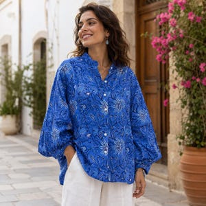Peut inclure: Une chemise bleue à boutons avec un motif floral. La chemise a des manches longues et un col officier. Le modèle porte un pantalon blanc. Le motif de la chemise comprend des accents blancs et noirs. La chemise est en tissu léger.