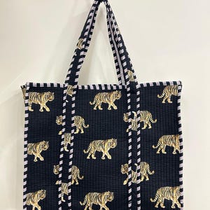 May include: Black tote bag featuring a repeating pattern of golden tigers. The bag has black and white striped handles and trim. The tote is rectangular and appears to be made of quilted fabric.
