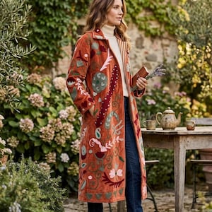 Könnte beinhalten: Ein langer, rostfarbener Mantel mit aufwendigen Stickereien mit Blumen- und abstrakten Mustern. Der Mantel hat einen Kragen und wird über einem cremefarbenen Rollkragenpullover und dunkelblauen Jeans getragen. Braune Lederstiefel vervollständigen das Outfit. Der Hintergrund ist ein Garten.