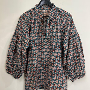 Op de afbeelding: Een blouse met lange mouwen in een blauwgroene stof met een herhalend patroon van kleine rode en crèmekleurige druppelvormen. Het kledingstuk heeft een hoge halslijn met een striksluiting en volumineuze mouwen die bij de polsen zijn geplooid.