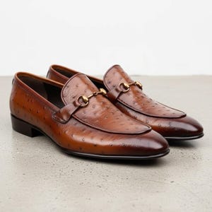 Könnte beinhalten: Ein Paar braune Leder-Loafer mit einer strukturierten Straußenleder-Optik und einem goldfarbenen Metall-Trensendetail über dem Vorderblatt. Die Schuhe haben einen niedrigen Absatz, eine abgerundete Spitze und ein poliertes Finish.