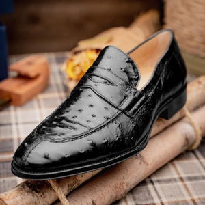 Könnte beinhalten: Ein schwarzer Slipper mit einer strukturierten Oberfläche und ausgeprägten Federfollikelmustern. Der Schlupfschuh verfügt über einen klassischen Penny-Steg über dem Vorderblatt, eine abgerundete Schuhspitze, einen flachen Absatz und ein glattes Innenfutter.
