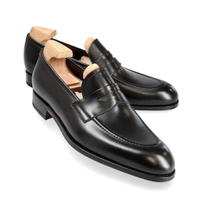 Puede incluir: Un par de mocasines penny de cuero negro con un diseño clásico de puntera tipo delantal y una tira horizontal sobre el empeine. Cada zapato incluye una horma de madera de color claro para mantener la forma de la piel superior.